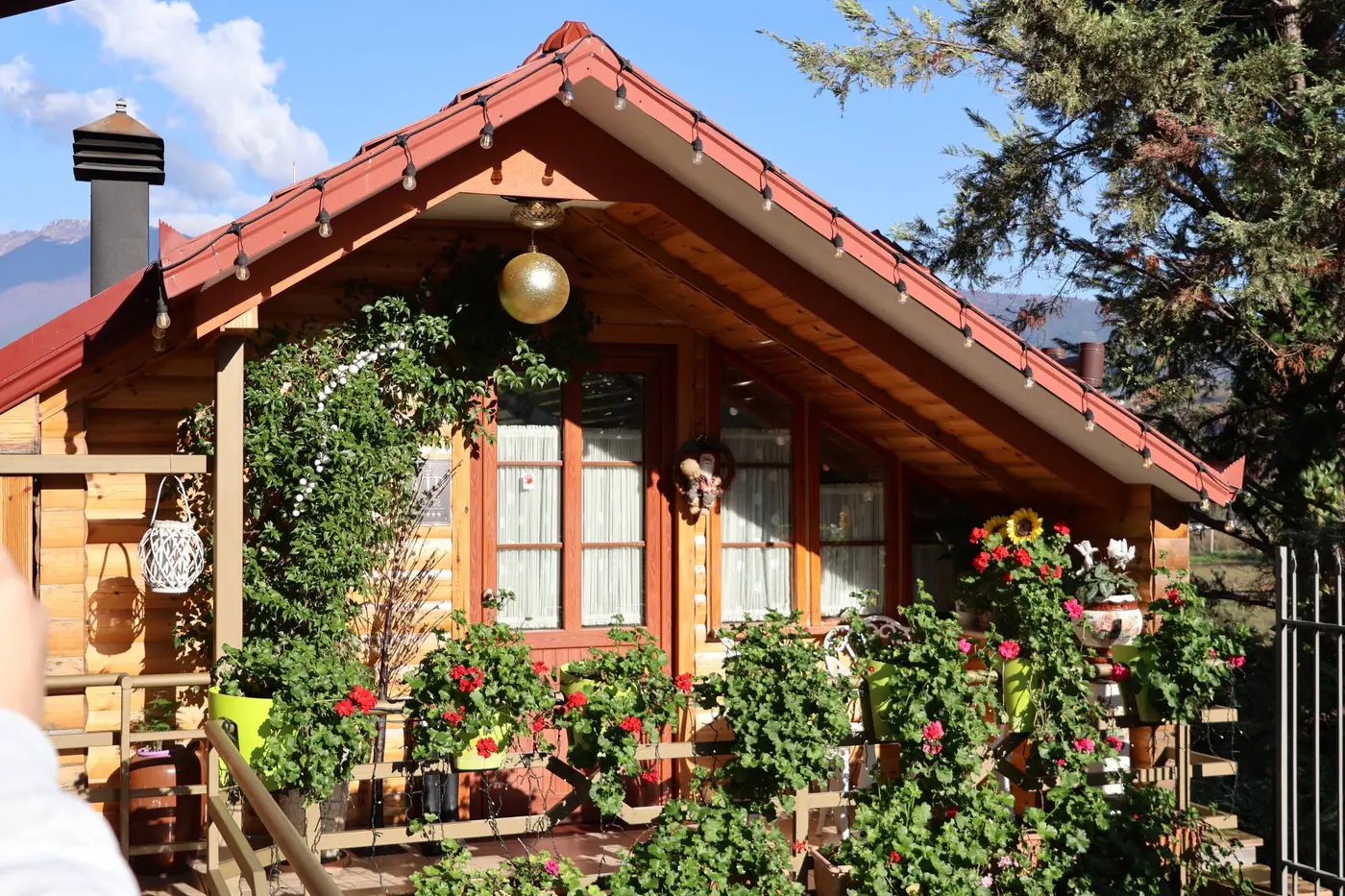 Veranda prej druri me lule në vazo, drita të varura dhe malet në sfond